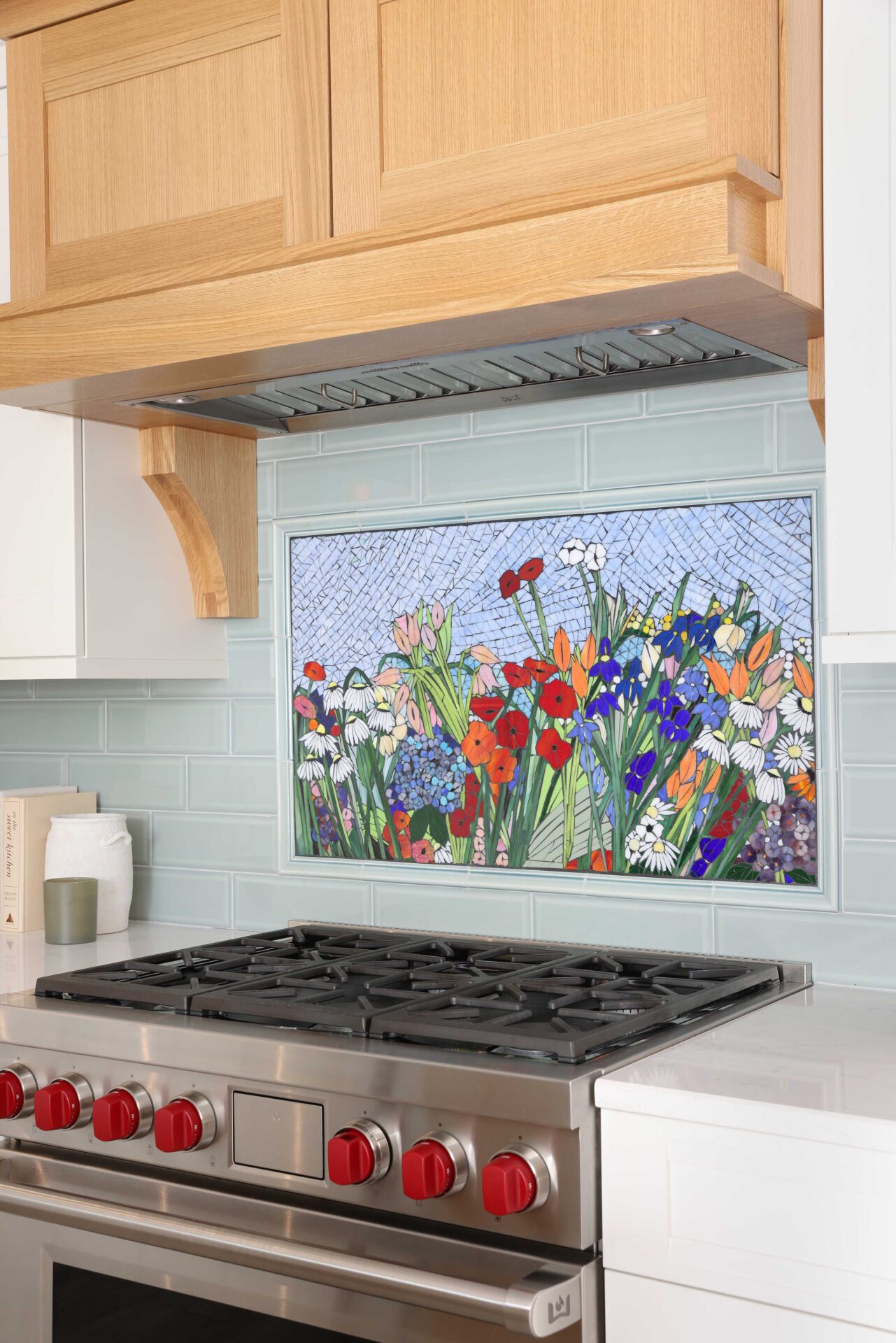 Colorful flower mosaic backsplash above range with surrounding light green subway tile, the bottom of a rift cut white oak range hood