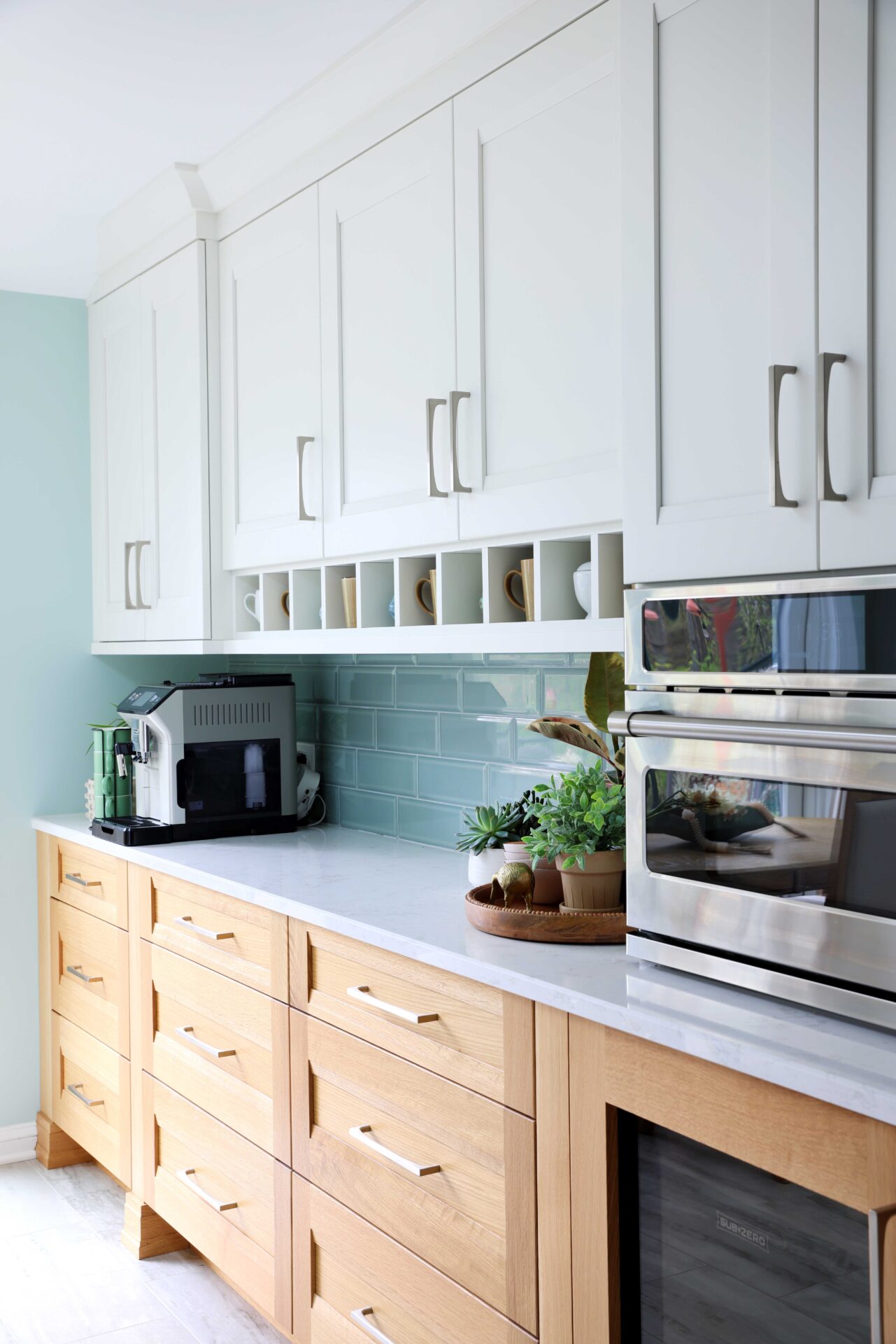 Upper white cabinets, rift cut white oak lower cabinets, coffee mugs in shelf nooks, light green tile backsplash, subway tile. cut white oak lower cabinets, coffee mugs in shelf nooks. Espresso machine on the left on counter. To the left, espresso machine on counter, to the right, stainless steel countertop microwave.