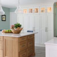 Rift cut white oak kitchen island with fruit on white countertop. White stacked cabinets with individual cabinets on top with glass panels and accent lighting with decorative elements inside, light green walls with art
