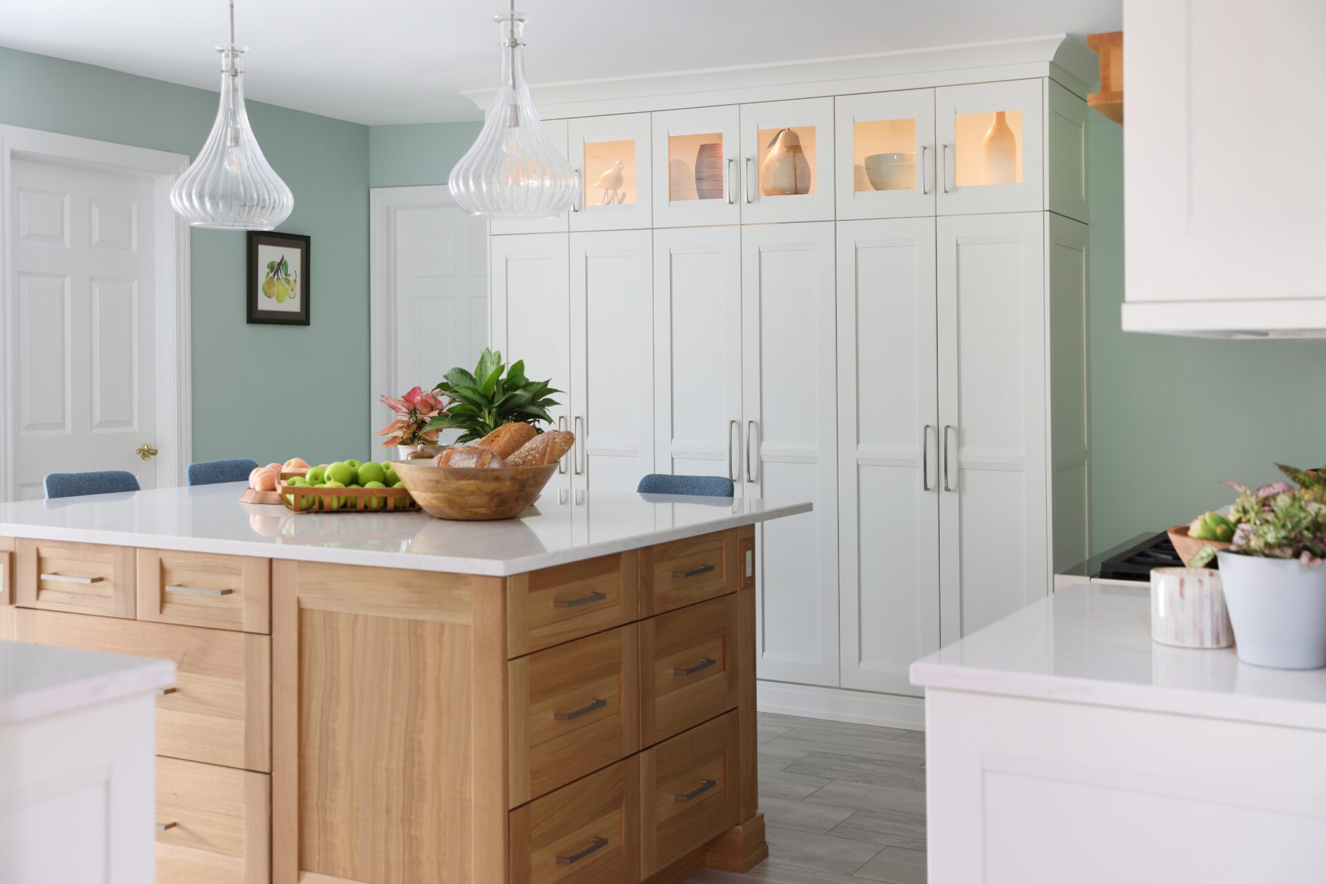 Rift cut white oak kitchen island with fruit on white countertop. White stacked cabinets with individual cabinets on top with glass panels and accent lighting with decorative elements inside, light green walls with art