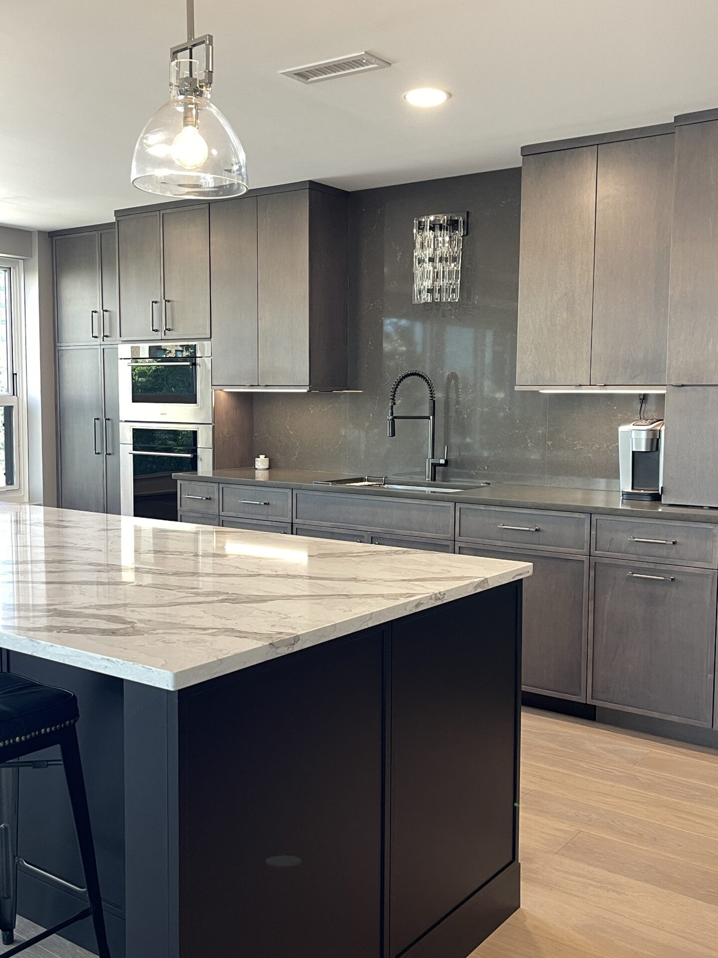 Black kitchen island with white countertop in foreground, dark brown maple cabinetry in background, with glass sconce above the sink