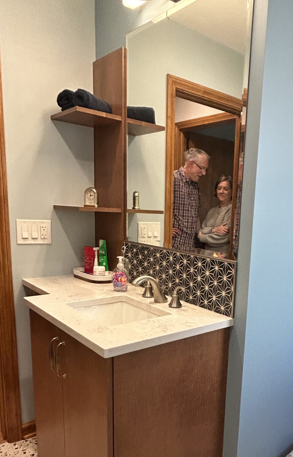 Wide shot of powder room with maple cabinetry, floating shelves, black rhomboid tile, reflection of homeowners in mirror