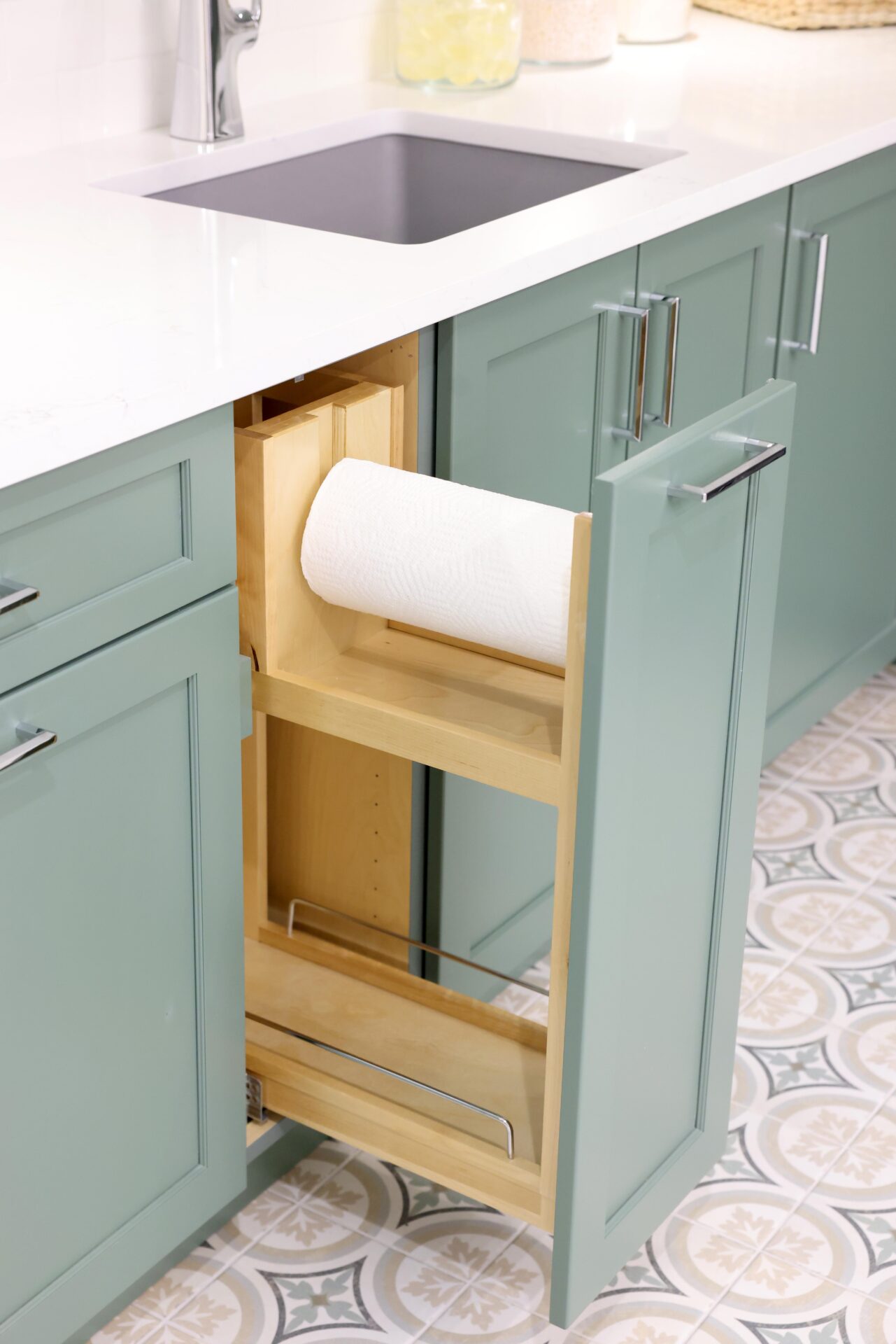 Green cabinets in laundry room with pull-out paper towel holder and storage shelf, white quartz countertops and gray sink