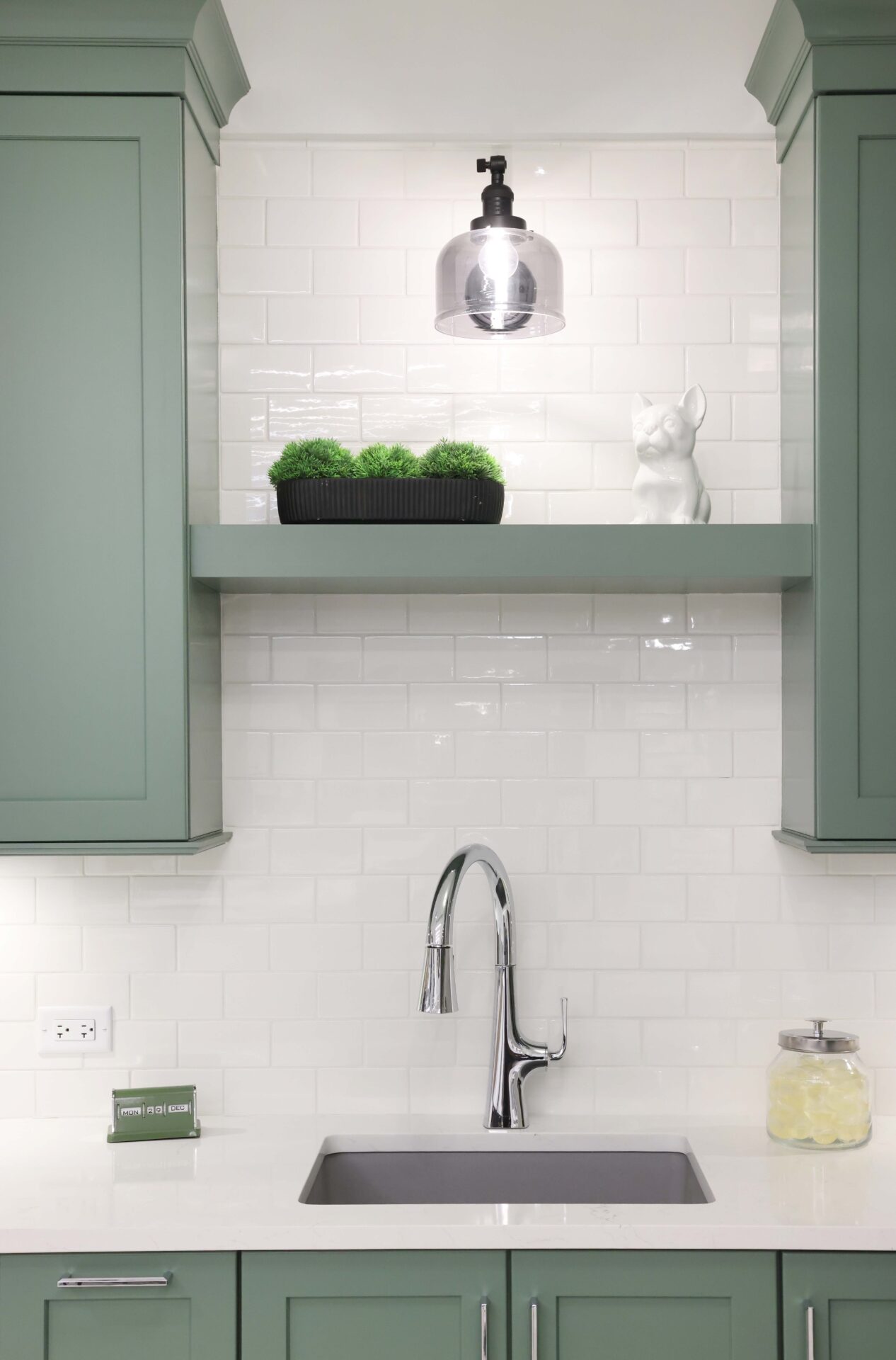 Green painted floating shelf in laundry room with green cabinetry, white subway tile backsplash, polished chrome faucet, white countertops, gray sink, matte black light fixture