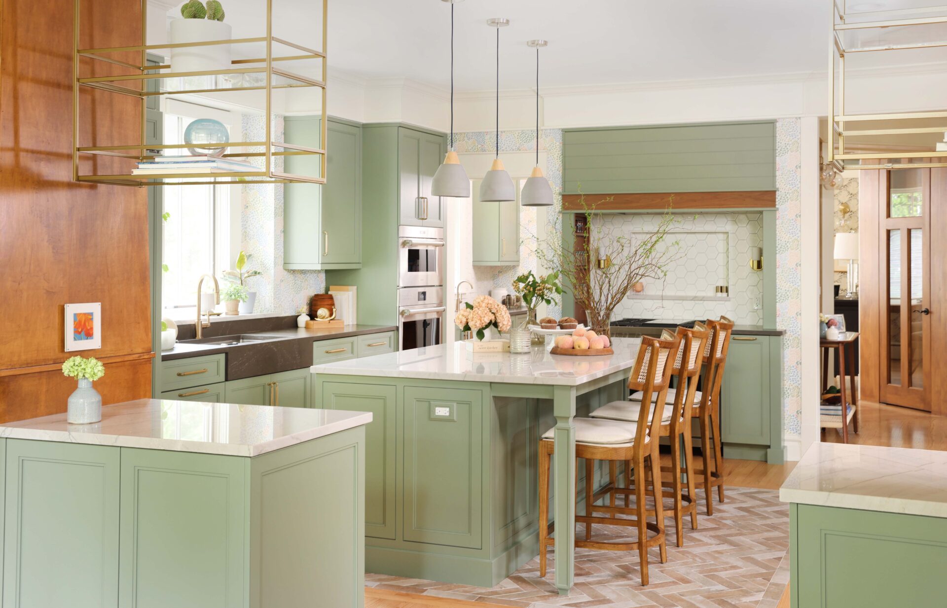 Natural stone white countertop at kitchen island, stacked stainless steel oven and microwave, light green cabinets, green hood with walnut wood trim, white geometric hexagonal backsplash with backsplash shelf and sconces on either side in range alcove with inset walnut spice shelves, suspended brass shelves, dark gray quartz countertop at sink area with counter apron style sink