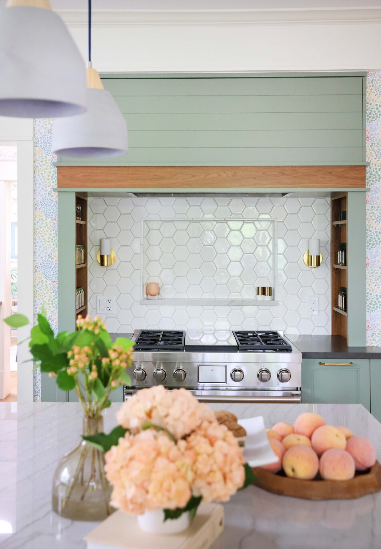 Green range hood with walnut wood trim, white geometric hexagonal tile backsplash with backsplash shelf, sconces