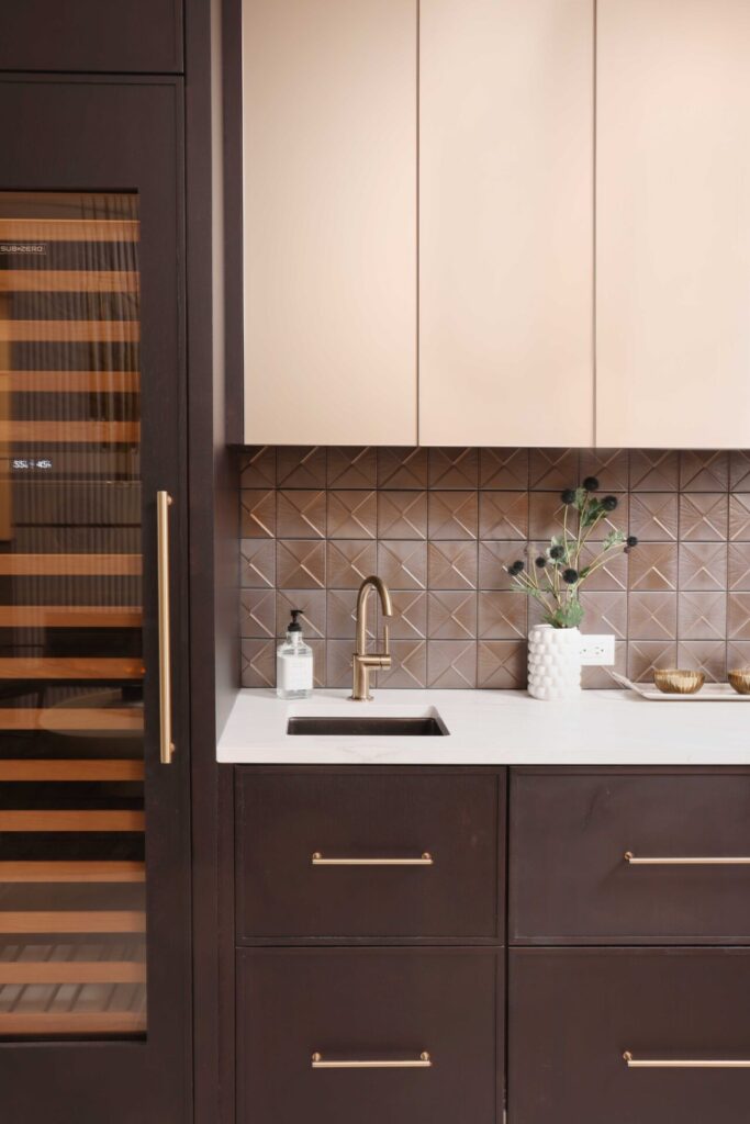 The modern beverage center features rift cut white oak cabinetry, ceramic tile backsplash, quartz countertops, wine fridge, integrated refrigerator drawers, brushed brass elements, and fireclay basin.