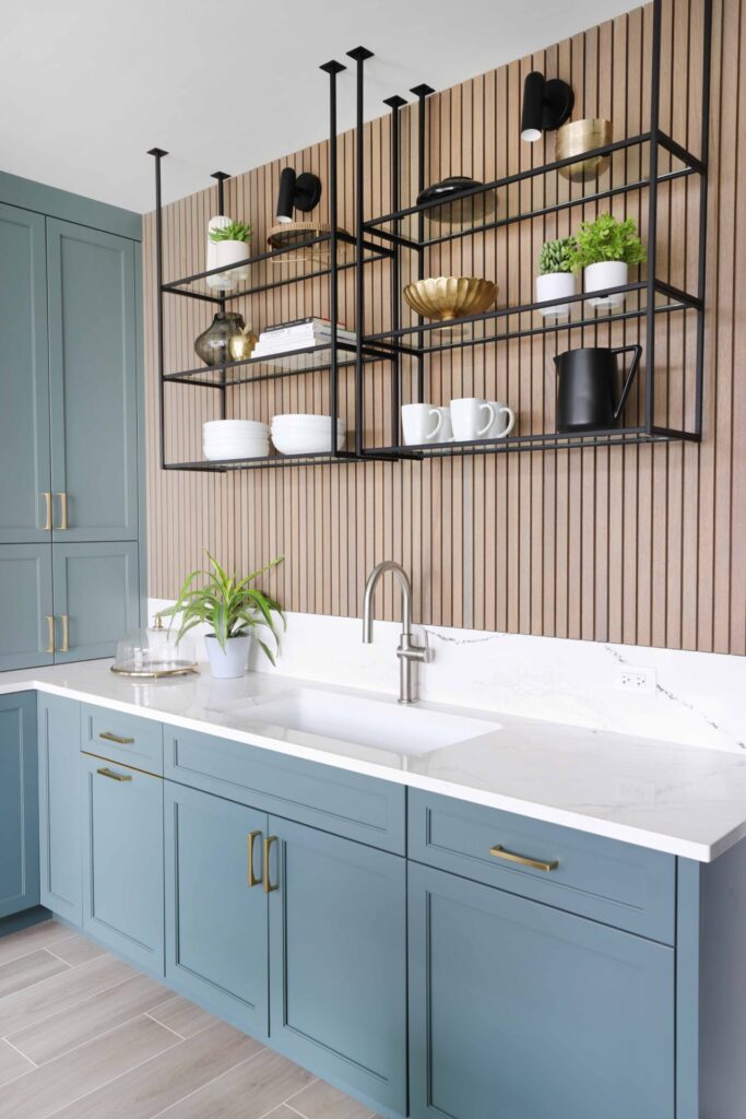 Above the spacious low-divide sink, matte-black metal shelves replace upper cabinets and offer more bold shapes, creating layered contrast in front of the oak slat paneling. Vertical wood slats at the sink wall and island are a rift cut white oak wood species that brings out beautiful color variations in stains.