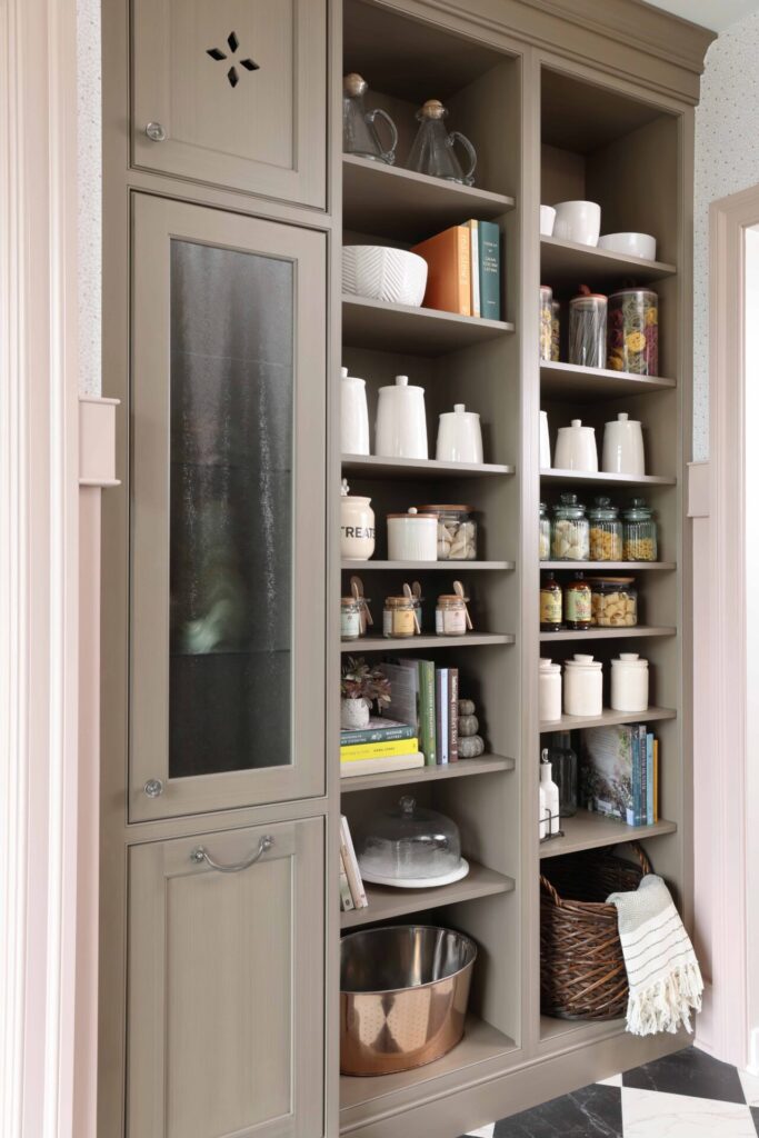 Garden estate butlers pantry features generous open shelving for everyday storage and display; custom cabinetry is painted in a soft, earthy mushroom tone with a brushed glaze and features vintage-style cutouts, furniture-like proportions, and tall baseboards that ground the space. Inside the cabinets are bottle holders, tip-outs for laundry or pet food, and thoughtful storage solutions tailored to daily life.