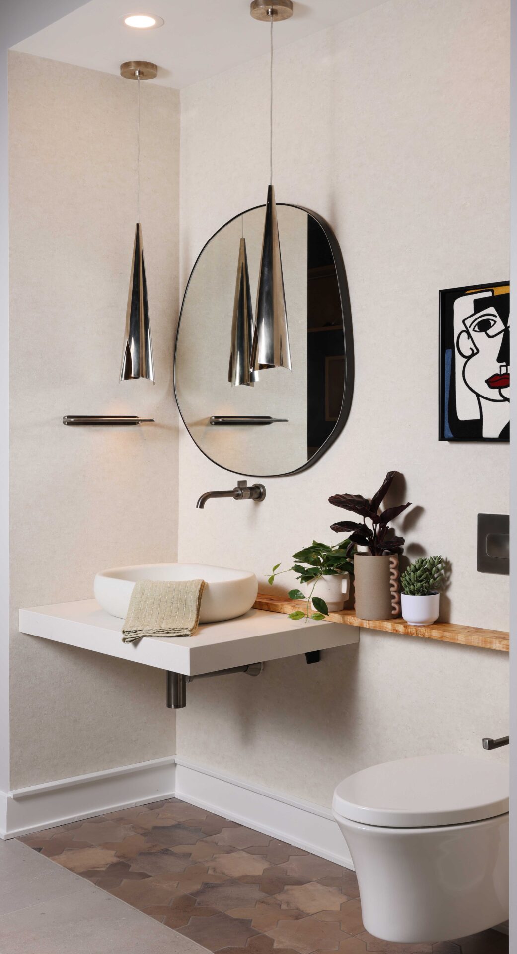 The Modern Mediterranean Powder Room features floating vanity and vessel sink designed to resemble rustic poured concrete; wall mounted faucet in luxe steel finish adding crisp architectural counterpoint. Plaster look wallpaper, burnished steel pendant lights, wall mounted toilet, blackened brass mirror, custom wood ledge, porcelain floor tile designed to mimic terracotta.