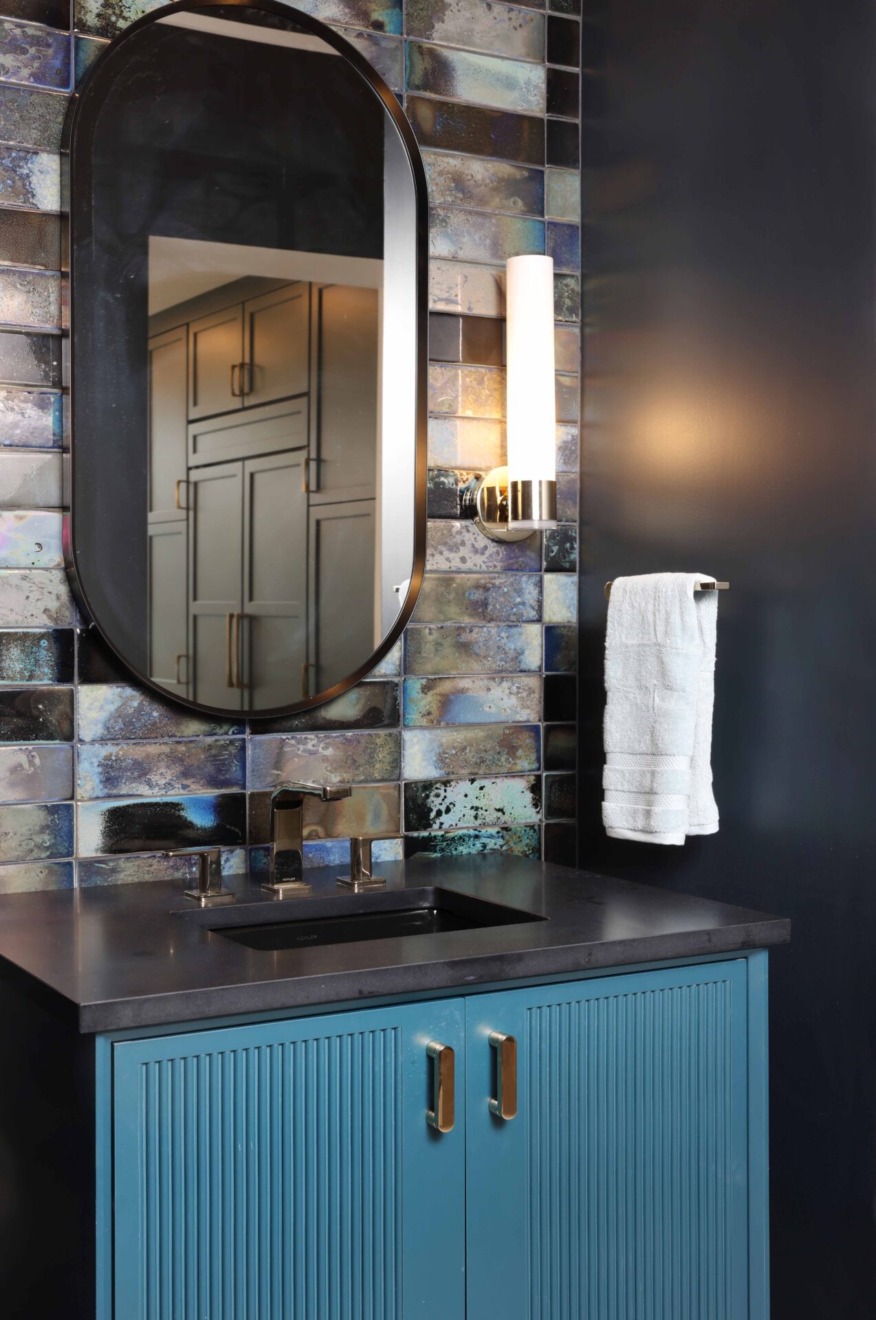 The dark and moody powder room features a custom teal vanity with reeded detailing, and iridescent tile paired with polished French gold hardware; custom legs complete the furniture-like feel, elevating the vanity from functional storage to statement piece. modern yet complementary gold sconces introduce a refined glow to the moody backdrop. Marble asymmetric herringbone floor adds classic pattern and subtle brightness underfoot.