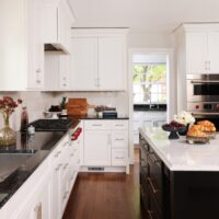 Modern kitchen design featuring white shaker cabinets, black countertops on left perimeter, a large island with black base and white countertop with gold hardware, stainless steel appliances, white paneled refrigerator natural wood flooring. A doorway peeks into a walk-in pantry with sink.