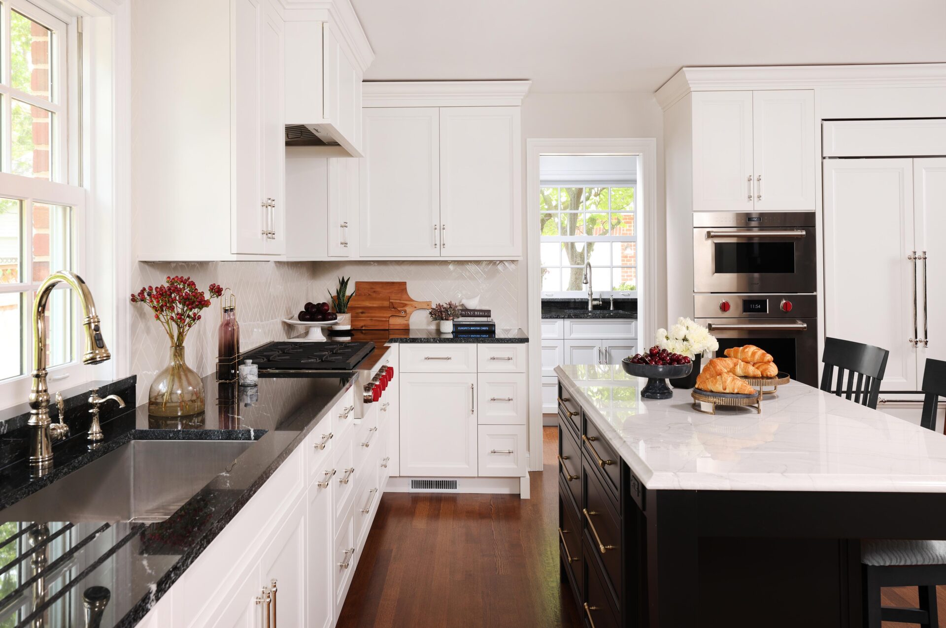 Modern white kitchen with island Modern kitchen design featuring white shaker cabinets, black countertops on left perimeter, a large island with black base and white countertop with gold hardware, stainless steel appliances, white paneled refrigerator natural wood flooring. A doorway peeks into a walk-in pantry with sink.