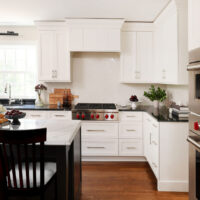 White range hood, white cabinets, range top with red knobs, black perimeter countertops, island with black base and white countertop. Window above sink. On the right stacked stainless steel ovens with red knobs
