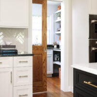 White kitchen cabinets with black countertops, wood pocket door half open showing a walk-in pantry with white cabinets, black countertops and sink with window above