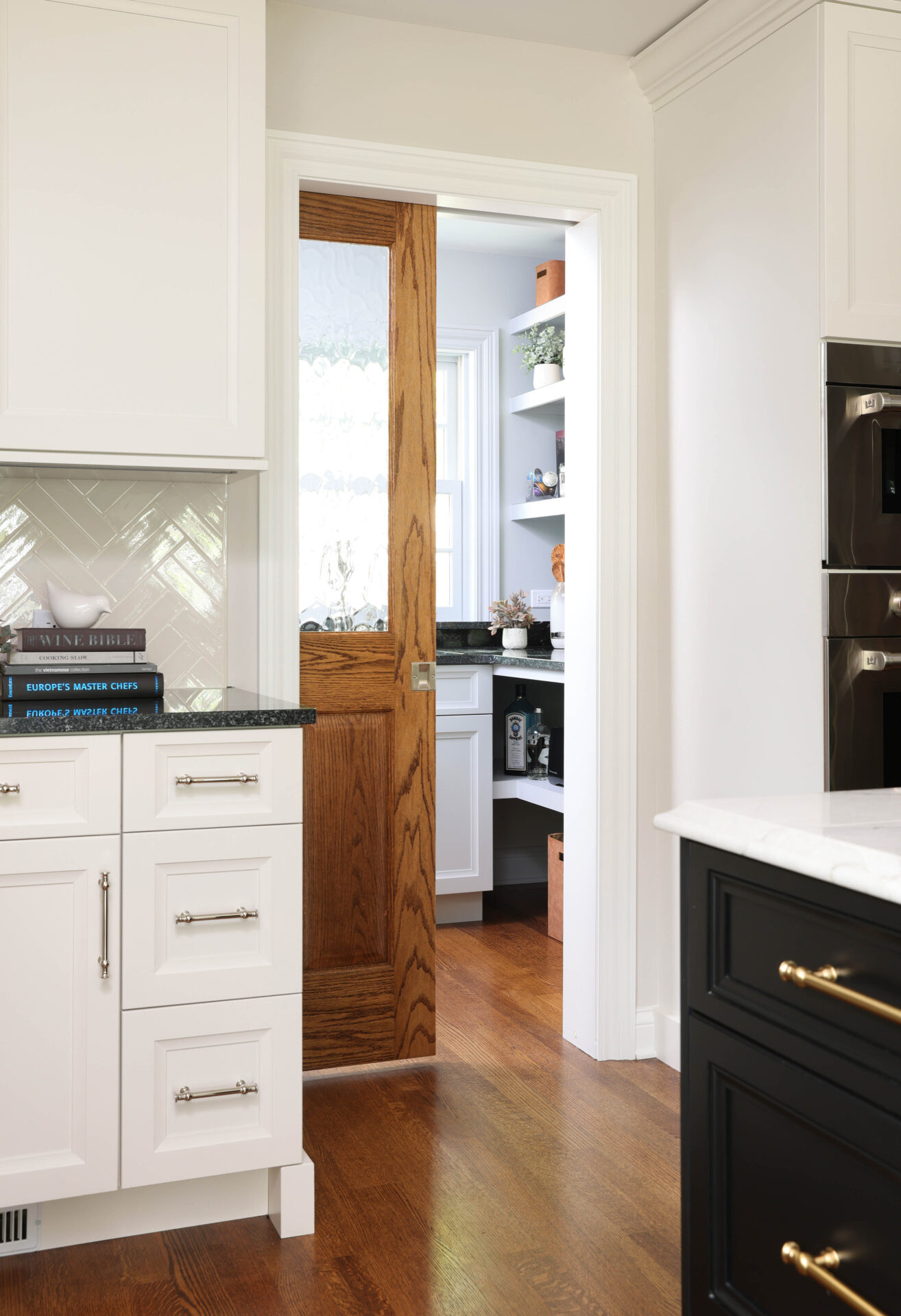 White kitchen cabinets with black countertops, wood pocket door half open showing a walk-in pantry with white cabinets, black countertops and sink with window above