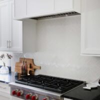 White herringbone backplash tile in kitchen, white range hood, range top with red knobs, white cabinets
