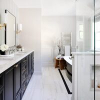 Elegant primary bathroom featuring black double vanity with quartzite white countertops on the left with two black-rimmed mirror above the two sinks and gold sconces with white lights. On the right is a black freestanding tub with white interior in the background near a stainless steel towel warmer. On the right in the foreground is the edge of a glass-enclosed shower with a white tile half wall and shower niche holding shampoo, conditioner and loofahs