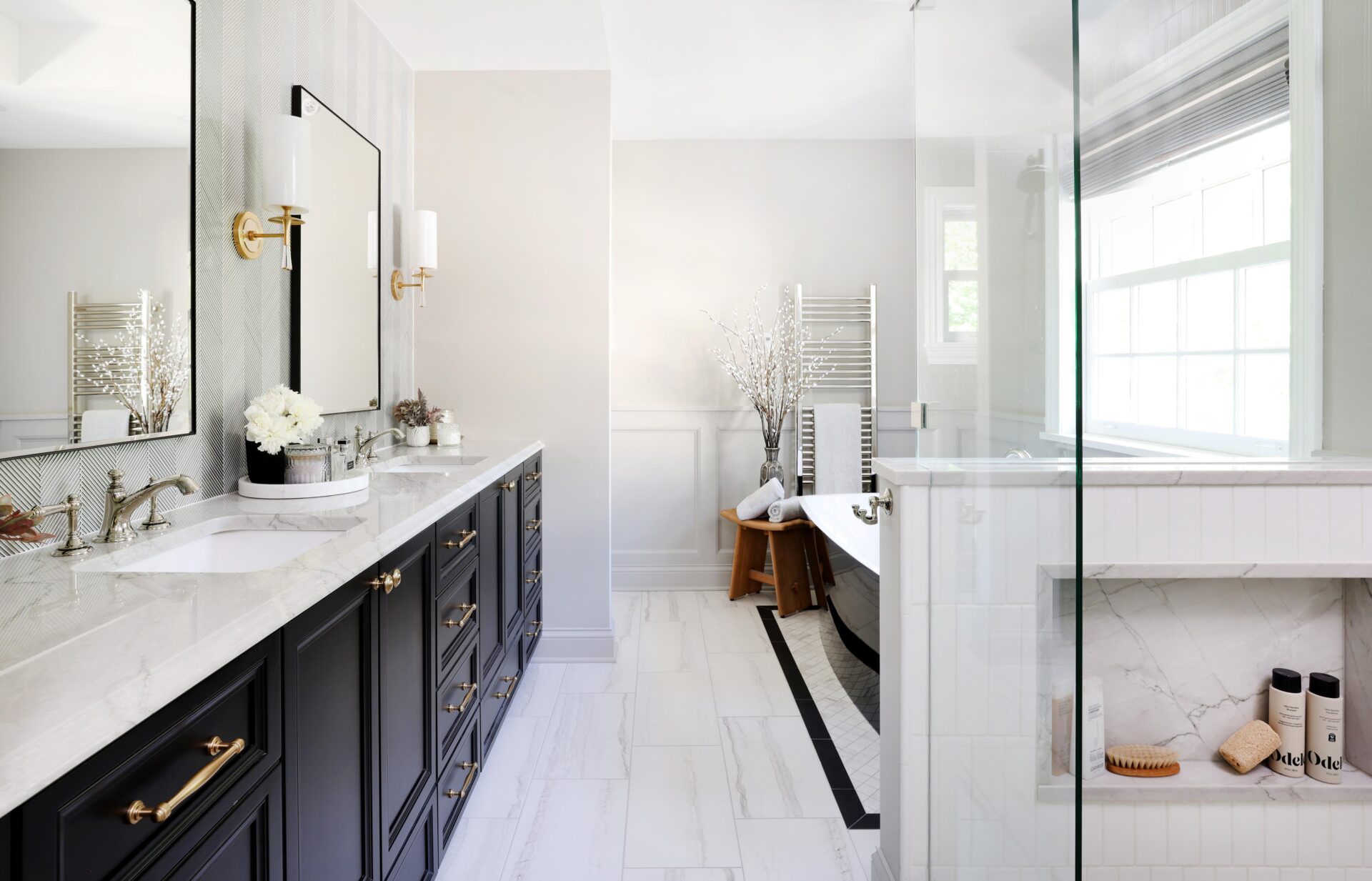 Primary bathroom with black double vanity and black tub Double black vanity with quartzite white countertops on the left with two black-rimmed mirror above the two sinks and gold sconces with white lights. On the right is a black freestanding tub with white interior in the background near a stainless steel towel warmer. On the right in the foreground is the edge of a glass-enclosed shower with a white tile half wall and shower niche holding shampoo, conditioner and loofahs