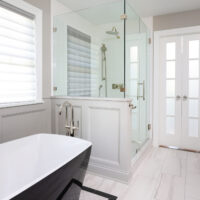 Freestanding tub with black exterior, white interior, polished nickel floor mounted faucet, white and black floor tile. Wainscoting half wall, glass shower with polished chrome rain shower head