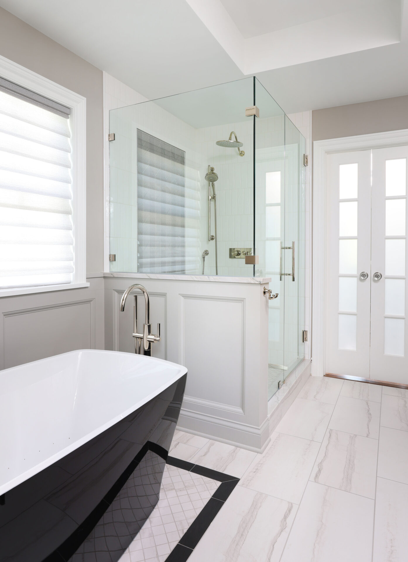 Black tub, glass shower, white bathroom Freestanding tub with black exterior, white interior, polished chrome floor mounted faucet, white and black floor tile. Wainscoting half wall, glass shower with polished nickel rain shower head